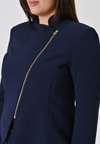 Navyblauw getailleerd jasje van een gladde stof met een hoge kraag en een schuine gouden ritsdetail aan de voorkant.