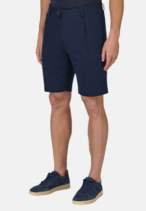 Un short bleu marine en tissu au toucher lisse, avec une fermeture à bouton, des passants de ceinture et des poches latérales. Assorti à des baskets bleu marine.