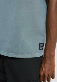 Lichtblauw T-shirt met korte mouwen van zacht materiaal, voorzien van een zwart logo label aan de zoom en een eenvoudige, gladde textuur.