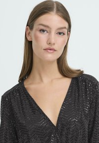 Czarna bluzka z cekinową fakturą, głębokim dekoltem w kształcie litery V i marszczonymi rękawami. Zdobiona błyszczącymi cekinami w różnych rozmiarach, tworzącymi kropkowany wzór.