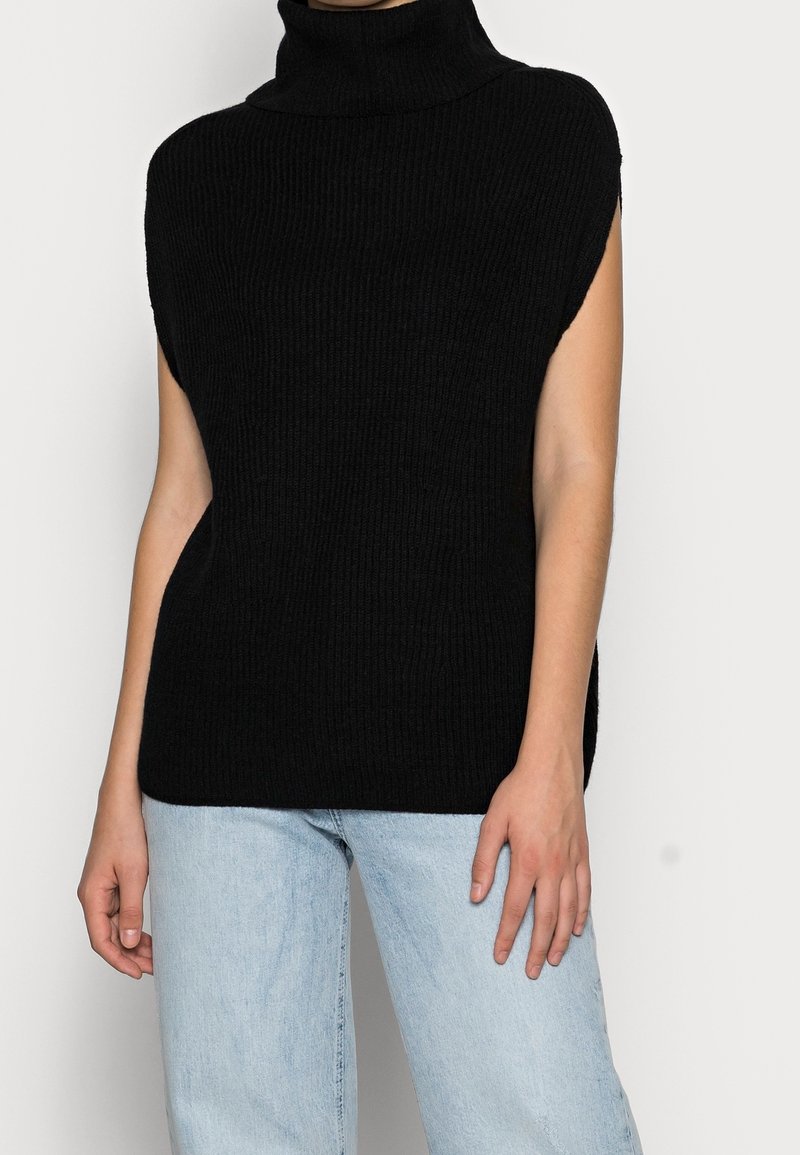 Haut à col roulé en maille noire avec manches courtes et texture côtelée, associé à un jean bleu clair. Design simple, sans motifs ni accents.