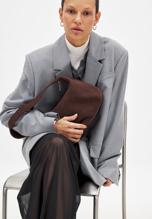 Bruine stoffen schoudertas met een gekromde vorm en ritssluiting, tegen een grijze oversized blazer en een doorschijnende zwarte rok gehouden.