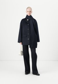 Marineblauer Nadelstreifenmantel mit großen Knöpfen und seitlichen Taschen, getragen über einem dunklen Outfit und kombiniert mit einer kleinen weißen Handtasche. Glatte Textur.
