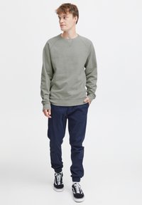 Sudadera gris con mangas raglán, cuello redondo, llevada con pantalones de chándal azul marino y zapatillas negras. Diseño sencillo, material de mezcla de algodón, ajuste casual.