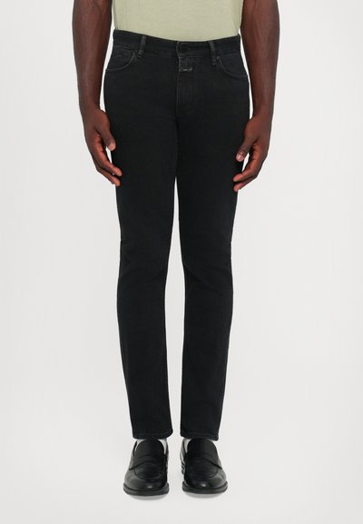 Homme portant un jean noir slim, des mocassins en cuir noir et une chemise jaune clair, se tenant devant un fond blanc uni.