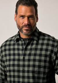 Camicia flanella a quadri verdi e neri con un design a bottoni, dotata di una tasca sul petto, tessuto strutturato e bottoni neri.
