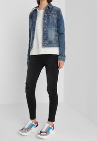 Denim jacket with metal button details, white knit sweater, black skinny jeans with distressed knees, and multicolored chunky sneakers.