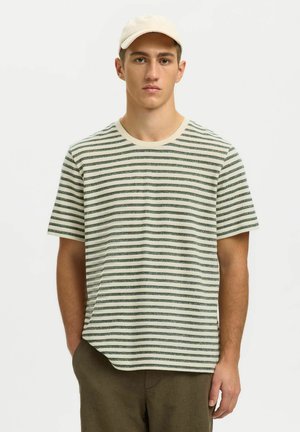 Jeune homme portant une casquette blanche, une chemise à manches courtes rayée verte et blanche, et un pantalon marron, debout avec une main dans la poche.