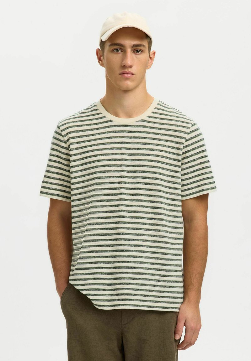 Młody mężczyzna w białej czapce, zielono-biały pasiasty krótki rękaw i brązowe spodnie, stojący z jedną ręką w kieszeni.