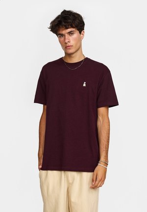 Jonge man met kort krullend donker haar die een donker bordeauxrode T-shirt en lichtbeige broek draagt tegen een effen achtergrond.