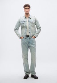 Ljusblå denimjacka med knäppning fram och två bröstfickor, kombinerad med något mörkare blå, löstsittande denimjeans och svarta skor.