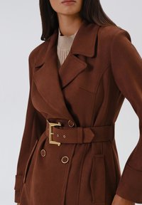 Trench coat marrone con cintura in tessuto morbido, ampi rever, dettagli in metallo dorato e bottoni doppi. Maglione a maglia testurizzata sotto.