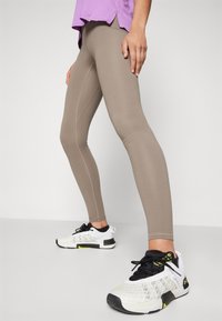 Beige leggings sima textúrával és varrás részletekkel, fekete-fehér sportcipőkkel párosítva, amelyek hálós felsőrésszel és sárga részletekkel rendelkeznek.