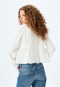 Witte peplum blouse van textuurstof, met lange mouwen en een gerende rand, gecombineerd met blauwe denim jeans met achterzakken.