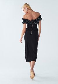 Black off-the-shoulder dress with ruffled neckline, bodycon shape, mid-length hem, and rear zipper detail. Worn with nude high heels.