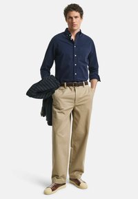 Marineblauwe button-up shirt, tan kleurige pantalon, bruine riem en bicolor sneakers. Een donkergestreepte blazer hangt over de arm.