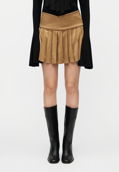 Jupe plissée en daim marron avec une taille ajustée, associée à des bottes en cuir noir montant jusqu'au genou. La jupe présente plusieurs plis verticaux.