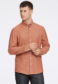 Camicia button-up color ruggine con maniche lunghe, bottoni bianchi, texture sottile e orlo curvato, mostrata su un modello maschile.