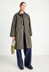 Woman wearing oversized olive green coat, wide-leg dark blue jeans, black shoes, holding olive green handbag against a plain background.