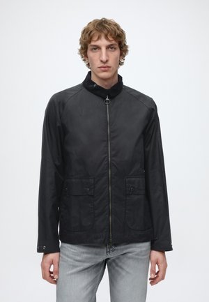 Junger Mann mit hellbraunem lockigem Haar, der eine schwarze Zip-Up-Jacke und hellgraue Jeans trägt und vor einem schlichten weißen Hintergrund steht.