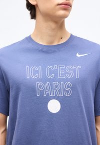 Personne portant un t-shirt Nike bleu avec le texte "ICI C'EST PARIS" et le logo du Paris Saint-Germain sur la poitrine, cadré au niveau du cou et des épaules.