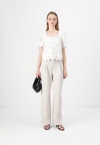 White lace blouse with scalloped hem, short puff sleeves, and button closure, paired with beige wide-leg trousers and a black bag.