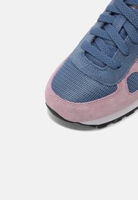 Gros plan sur la pointe d'une basket bleue et rose avec tissu en mesh, superpositions en daim et lacets bleus sur un fond blanc.