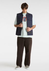 Gilet senza maniche blu navy con due tasche frontali su una t-shirt verde menta con stampa grafica, abbinato a pantaloni marroni e scarpe slip-on a scacchi.
