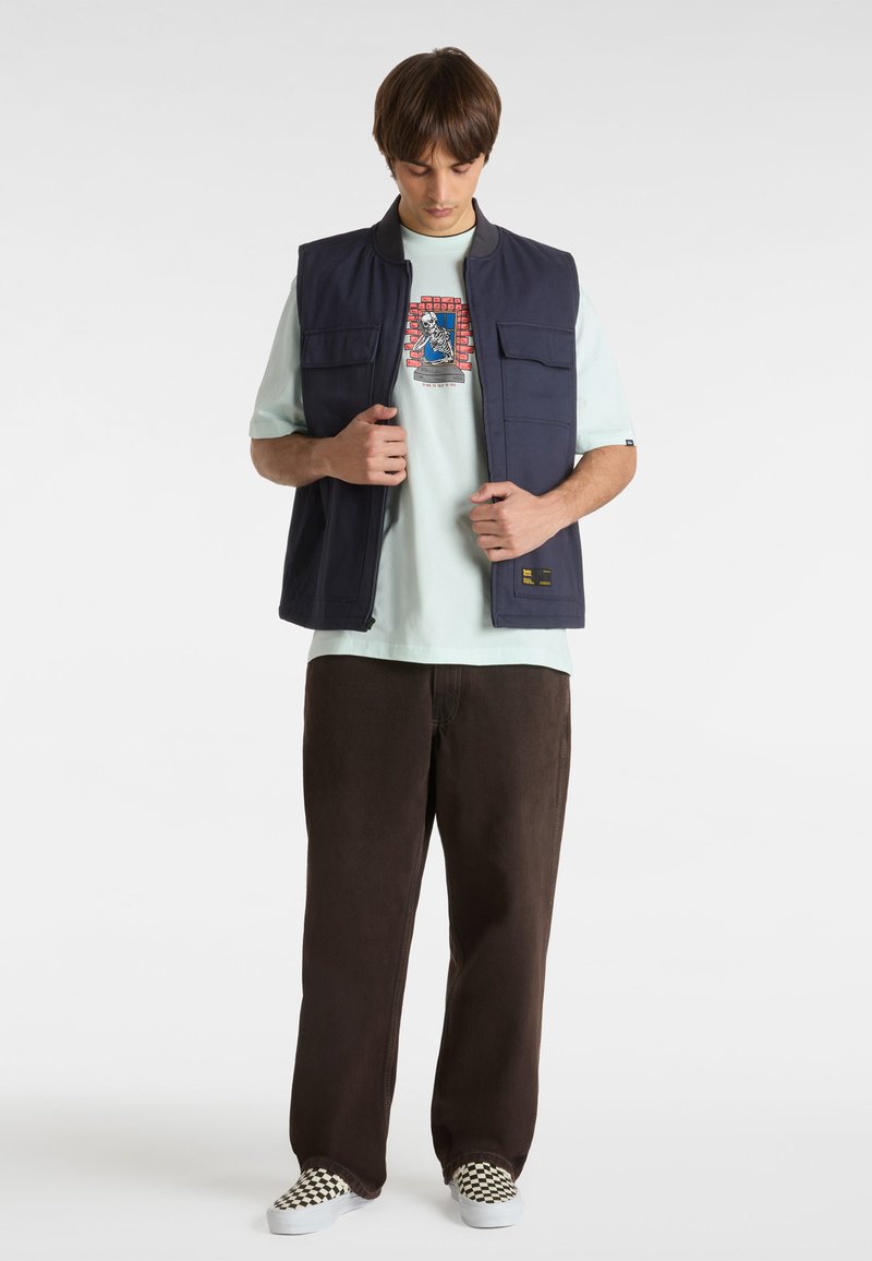 Chaleco sin mangas azul marino con dos bolsillos frontales sobre una camiseta verde menta con un estampado gráfico, combinado con pantalones marrones y zapatos de cuadros tipo slip-on.