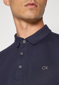 Námornícky modrá pižamo s textúrovanou tkaninou, dvoma gombíkmi pri golieri a malým bielym vyšitým logom "CK" na hrudi.