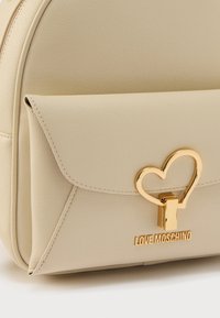Μπεζ τσάντα από συνθετικό υλικό με υφή και χρυσό κούμπωμα σε σχήμα καρδιάς. Διαθέτει μπροστινή τσέπη και ανάγλυφο λογότυπο "LOVE MOSCHINO".