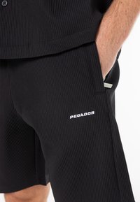 Homme portant un short côtelé noir avec la main dans la poche, montrant le logo blanc "PEGADOR" sur la cuisse.
