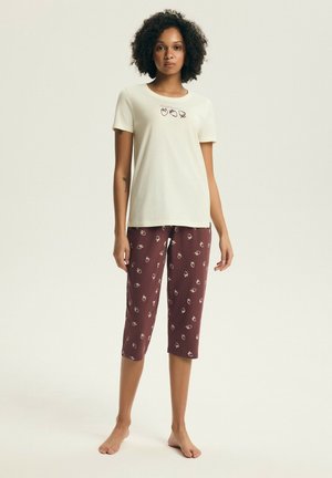 Kobieta w kremowej bluzce z krótkim rękawem ozdobionej małymi grafikami owoców oraz w bordowych, skróconych spodniach z dopasowanym wzorem owoców, stojąca boso.