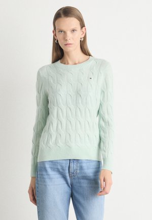 Light mint green cable-knit sweater with ribbed cuffs and hem, featuring a small brand logo on the chest. Paired with blue jeans.