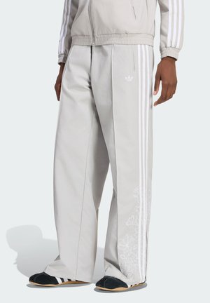 Muž nosící světle šedé tepláky Adidas s bílými pruhy a květinovým vzorem podél nohou, doplněné o odpovídající bundu a černé boty.