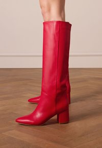 Red leather knee-high boots with a pointed toe and chunky block heel, featuring a smooth texture and minimalist design.