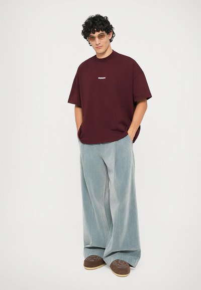 T-shirt oversize bordeaux avec le texte blanc "BONSAI", associé à un pantalon large bleu clair et des chaussures brunes à enfiler.