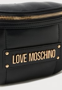 Juodasis dirbtinis odinis maišelis su auksiniu užtrauktuku. Yra išsiskirianti auksinė plokštelė su užrašu "LOVE MOSCHINO" ir dekoratyviniai kaiščiai.