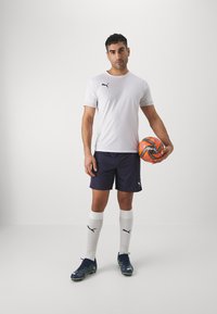 PUMA sportfelszerelés: fehér kereknyakú póló, sötétkék rövidnadrág, fehér zokni és sötét cipő. Narancssárga és fekete focilabda a jobb kezeiben.