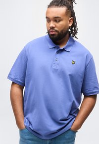 Άνδρας με κοτσιδωτά μαλλιά που φοράει μπλε πόλο πουκάμισο με κίτρινο λογότυπο πουλιού, με τα χέρια στις τσέπες, κοιτάζοντας προς τα κάτω και πλάγια, μπροστά σε απλό φόντο.