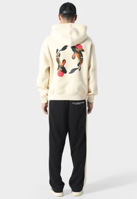 Crème hoodie met een koi vis grafiek op de achterkant en rode cirkels, gecombineerd met zwarte sweatpants met witte zijstrepen en een logo.