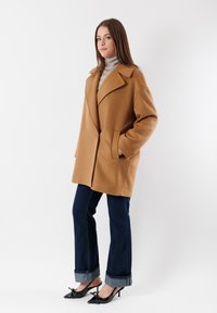 Cappotto oversize in misto lana marrone con ampi revers e tasche laterali; abbinato a jeans svasati in denim scuro e tacchi in pelle verniciata nera.