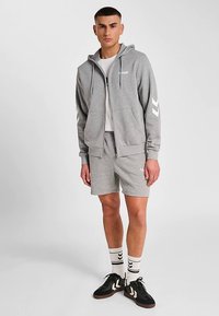 Lichtgrijze hoodie met rits en capuchon, korte grijze shorts en zwarte sneakers. Geaccentueerd met witte chevronpatronen en branding.
