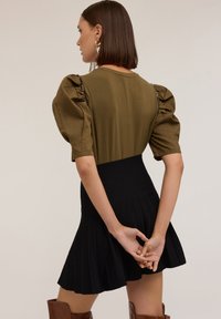 Top verde oliva con mangas abullonadas, adornado con detalles en negro, combinado con una falda negra de corte ajustado y plisada, que muestra una textura suave y un contraste.