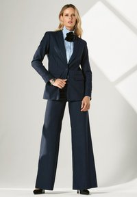 Marineblauw suit met tenue blazer en wijde broekspijpen. Bevat een lichtblauw shirt en zwarte bloemen halsaccessoire. Soepele textuur.