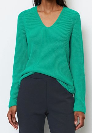 Femme portant un pull en laine vert à manches longues avec un col en V, rentré dans un pantalon noir, debout devant un fond clair uni.