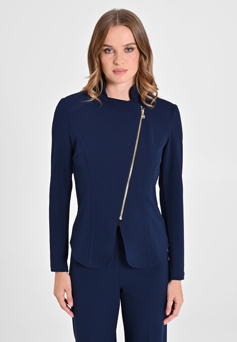 Blazer bleu marine à coupe ajustée, manches longues et fermeture éclair en diagonale dorée. Fabriqué en tissu lisse avec une silhouette épurée et minimaliste.