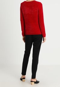 Červený svetr z texturovaného úpletu s dlouhými rukávy a kulatým výstřihem; kombinovaný s černými úzkými kalhotami a černými slip-on botami s dřevěnými podpatky.