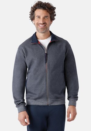 Mann mit lockigem Haar, der lächelt, trägt eine dunkelgraue Jacke mit Reißverschluss über einem weißen Hemd und dunklen Hosen, steht vor einem einfarbigen Hintergrund.
