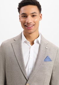 Lichtgrijze blazer met een gestructureerde weving, voorzien van een blauwe pochet, een wit shirt eronder en een reversspeld aan de linkerkant.
